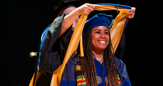 Smiling doctoral student in regalia being hooded on stage at Commencement